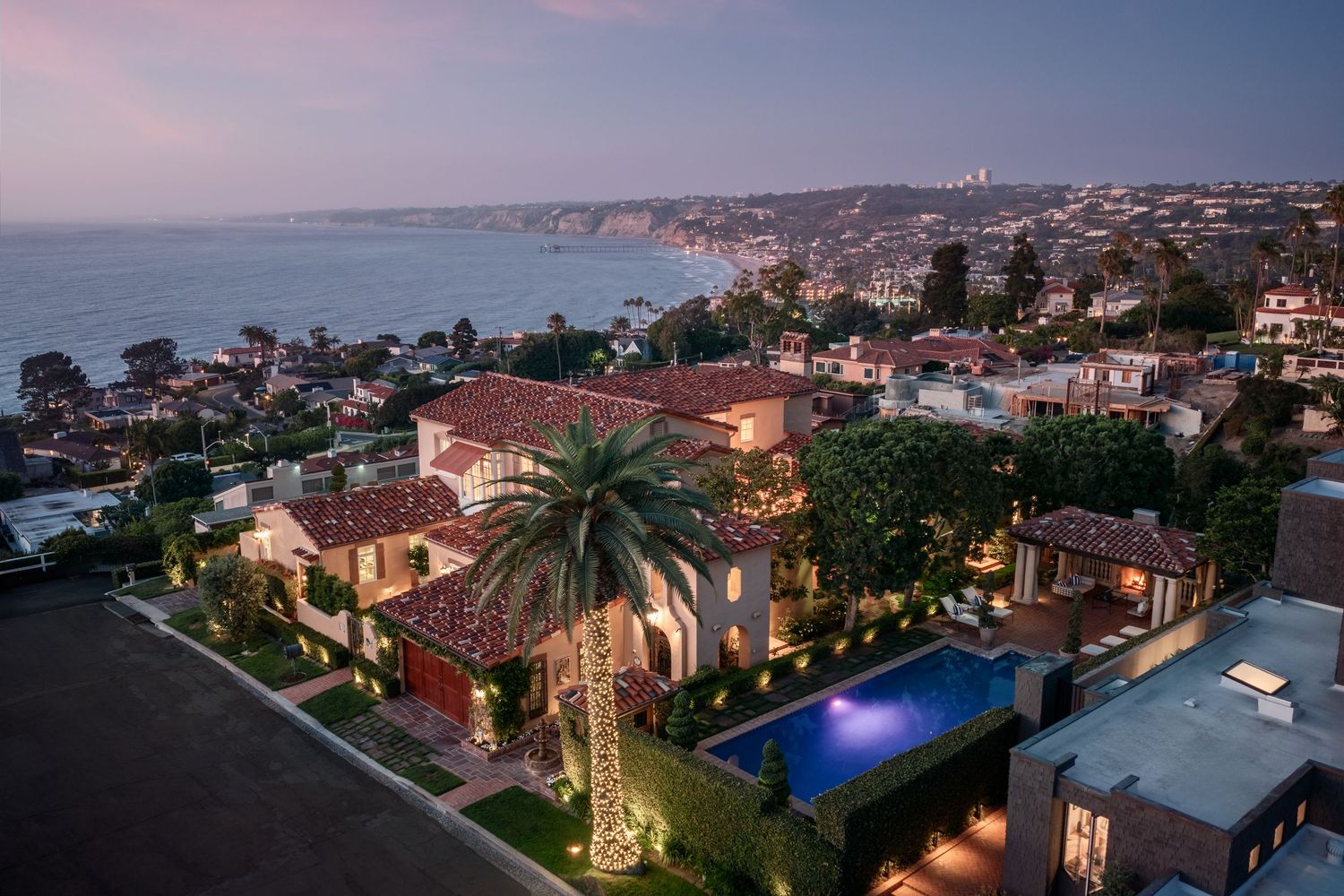 Aerial twilight view of a luxury La Jolla coastal home with ocean views, palm trees, pool, and hillside neighborhood at dusk.