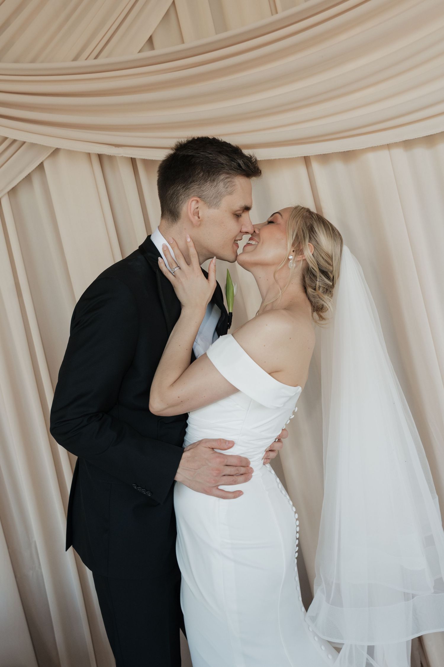 Bride and groom share a romantic kiss in front of elegant draped beige curtains at their wedding celebration.