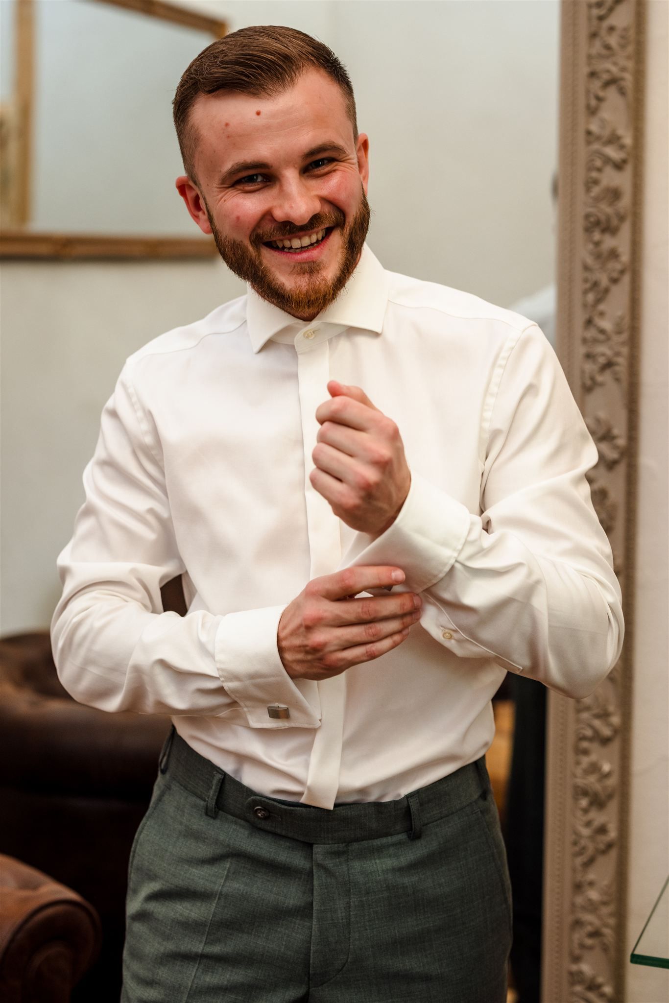 A person in a crisp white dress shirt and olive pants smiles while adjusting their cuff in an elegant interior setting.