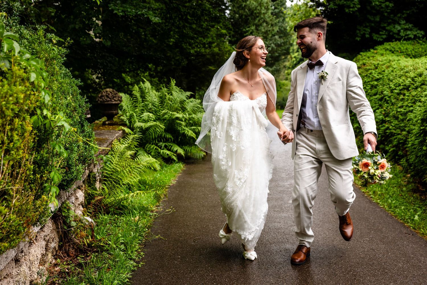 A couple in wedding attire walks together along a lush garden path holding hands.