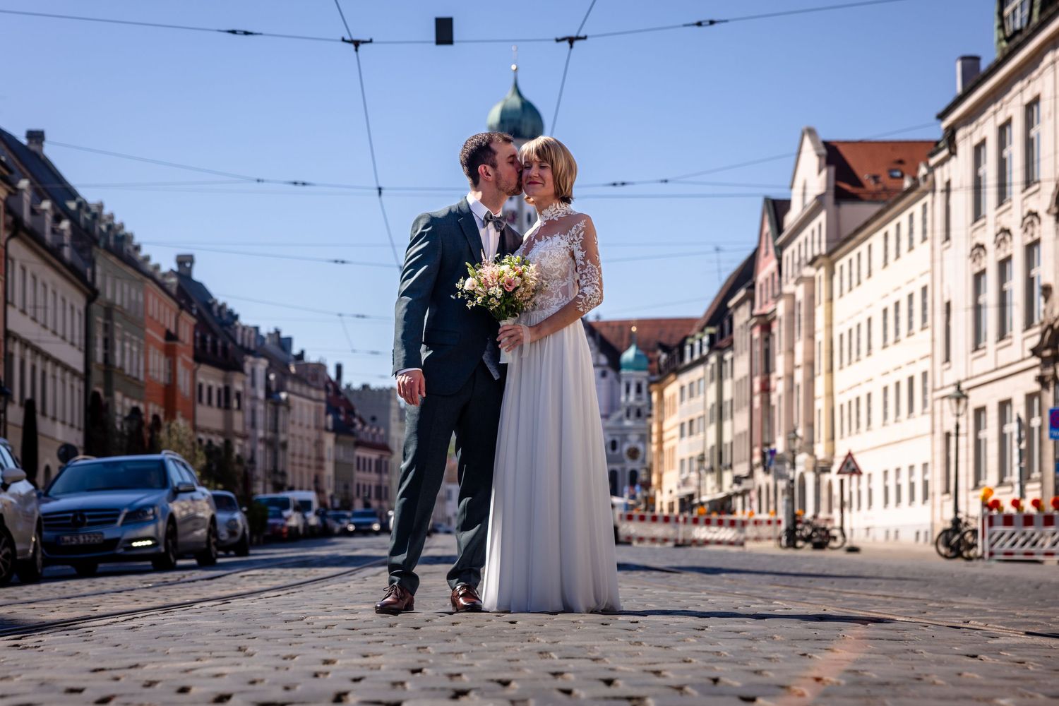 Bride and groom kissing on cobblestone street with colorful European buildings in sunny city backdrop.