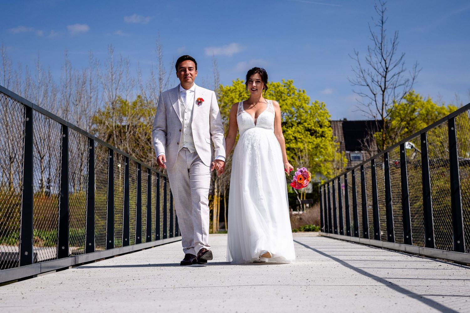 Bride and groom holding hands on a bridge outdoors on their wedding day, wearing white dress and suit.