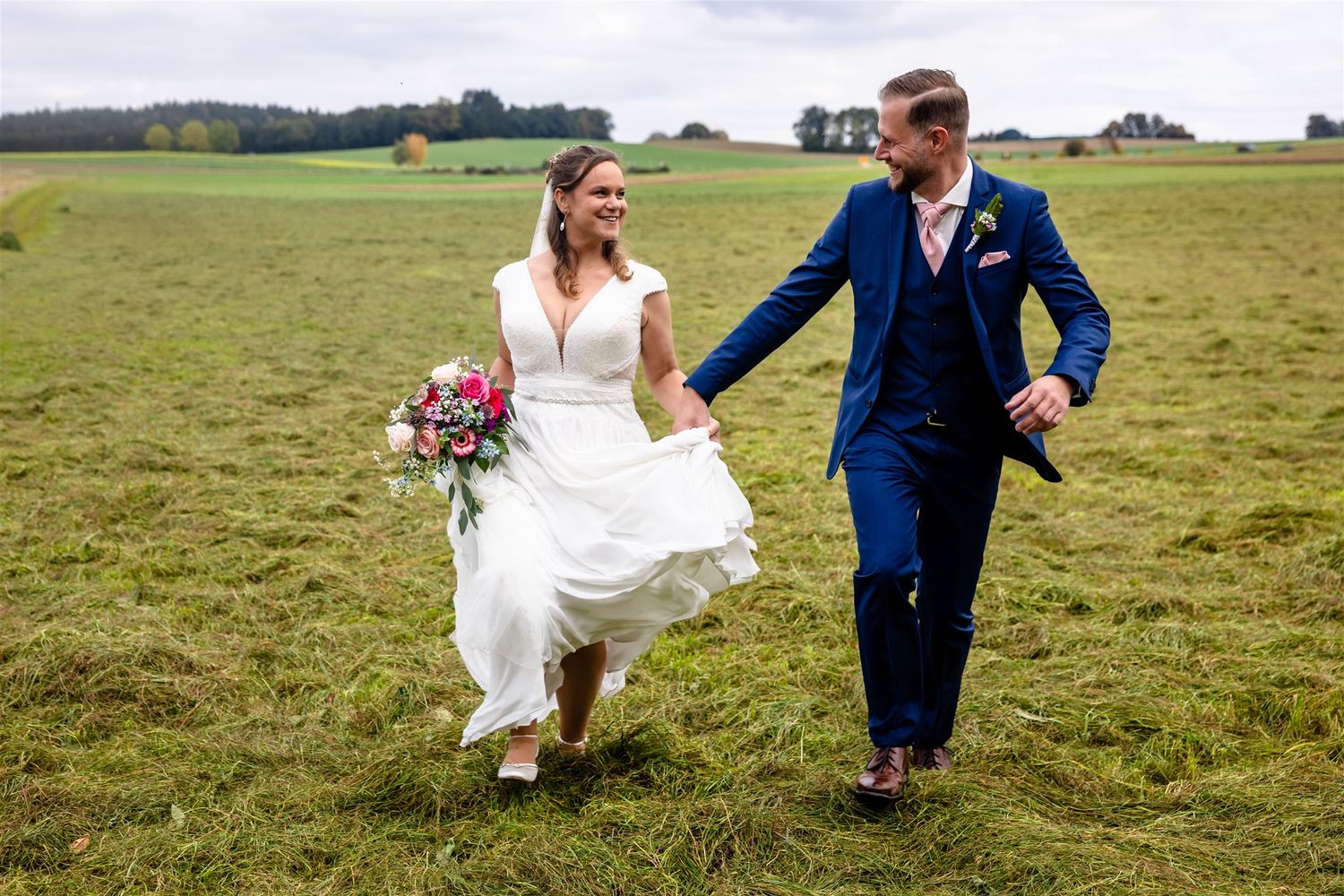 A couple in wedding attire playfully runs and jumps through a green grassy field on their special day.