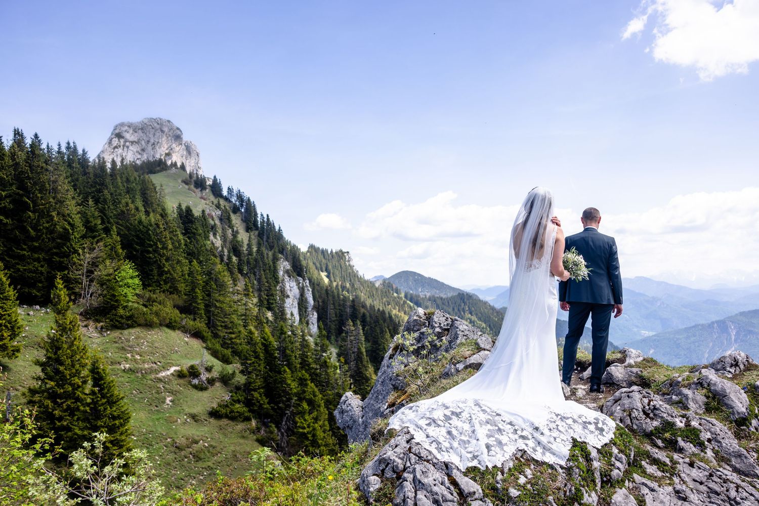 Bride and groom stand on rocky mountain summit overlooking lush alpine valley on their wedding day.