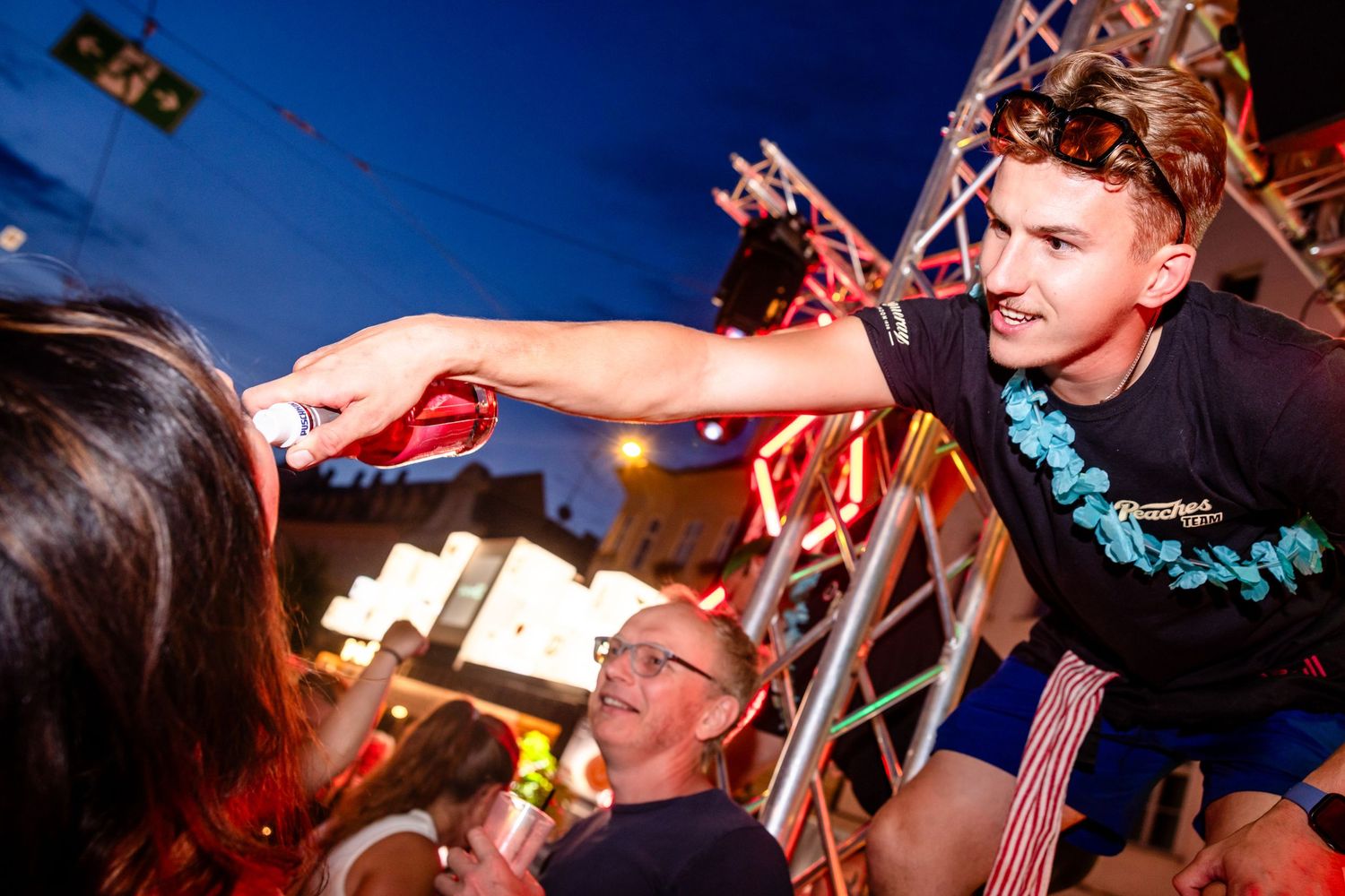 Young man in blue lei handing drink to crowd at outdoor night festival with colorful lights.