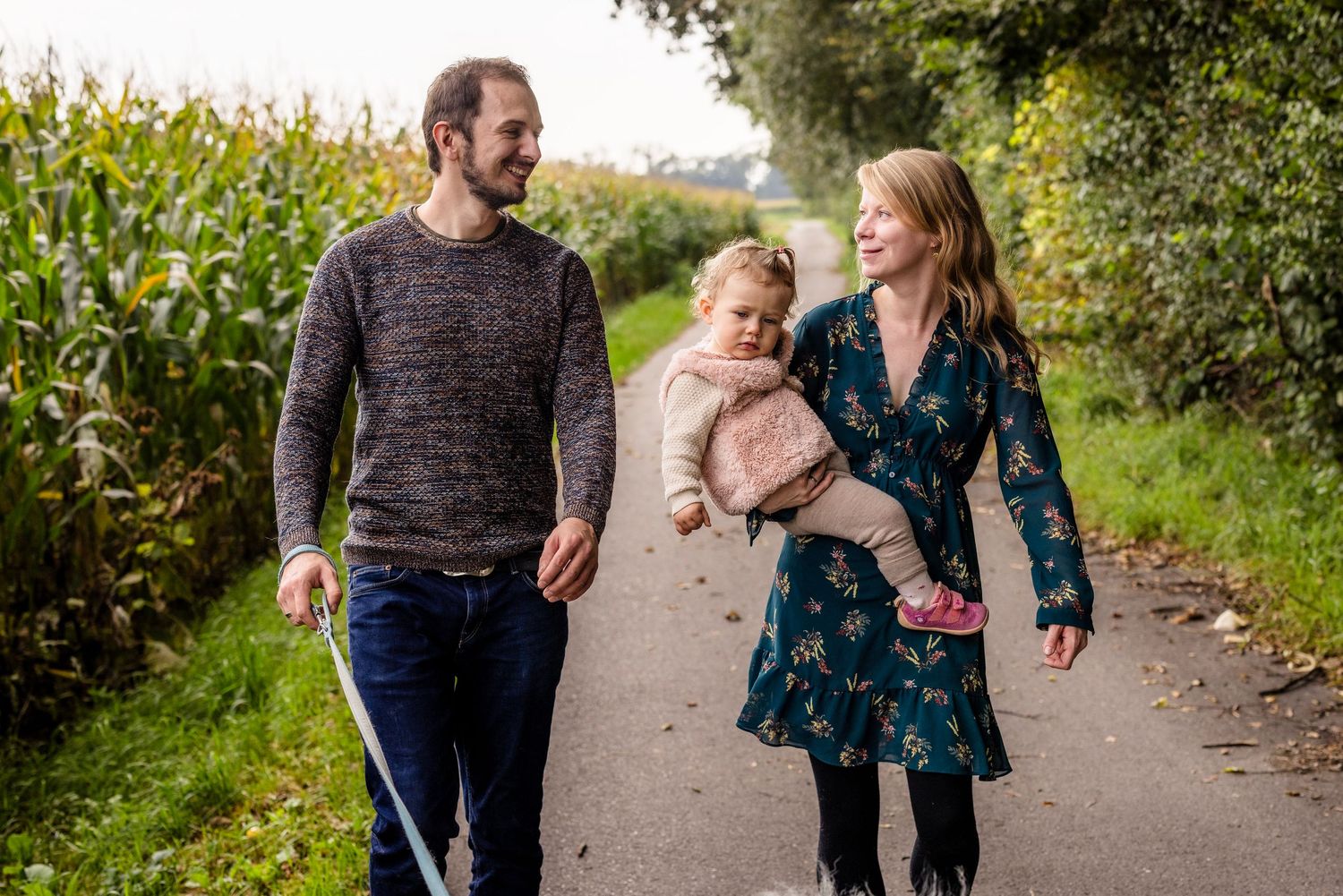 A family walks together and laughs on a path bordered by corn fields and greenery on a sunny day.