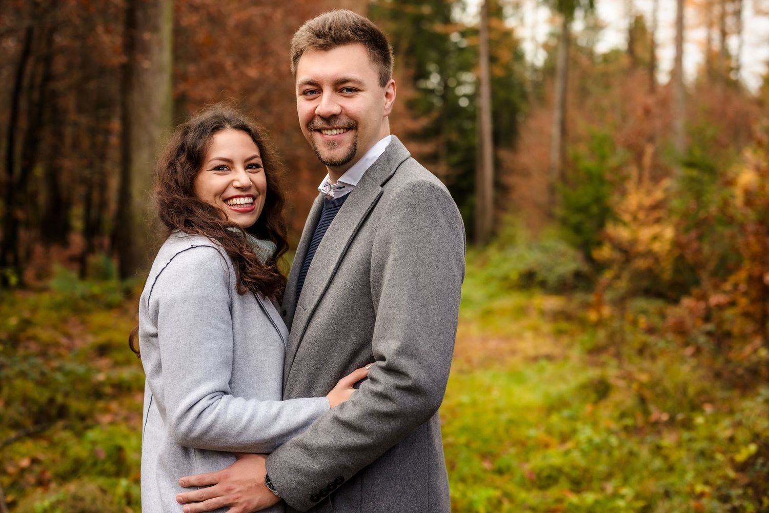 Smiling couple embracing in an autumn forest, wearing gray jackets, surrounded by colorful fall foliage.