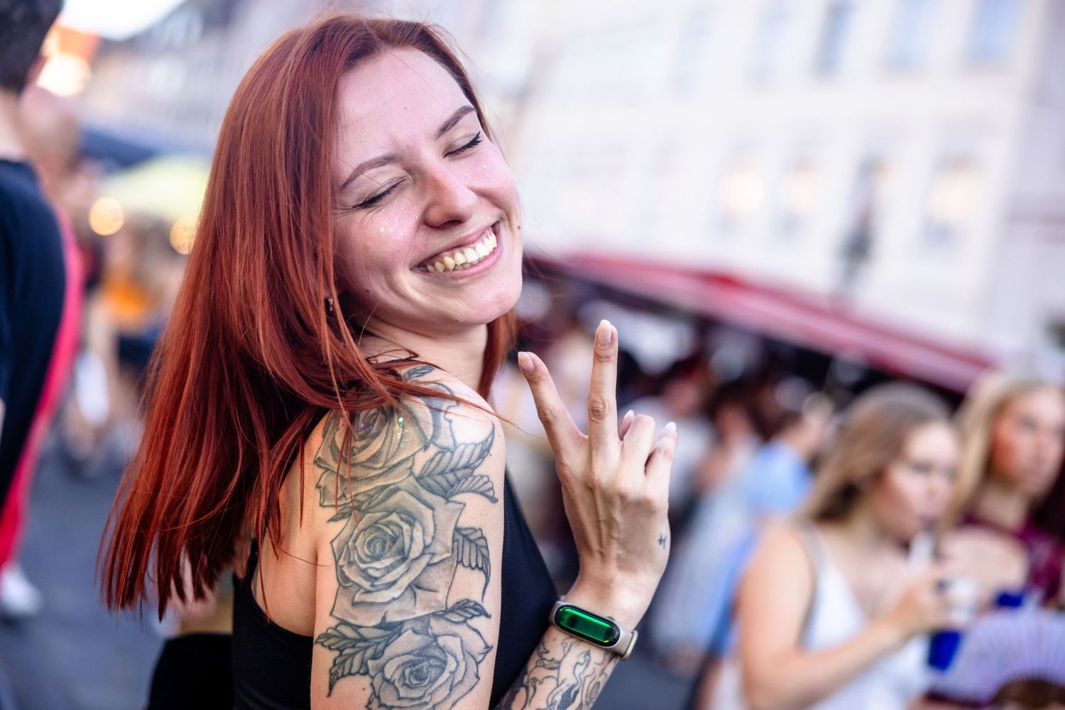 Smiling redhead woman with rose tattoos on her shoulder making a hand gesture at an outdoor street event.