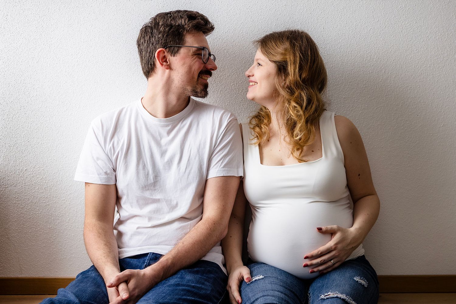 A couple in white shirts sits together against a white wall while smiling and looking at each other.
