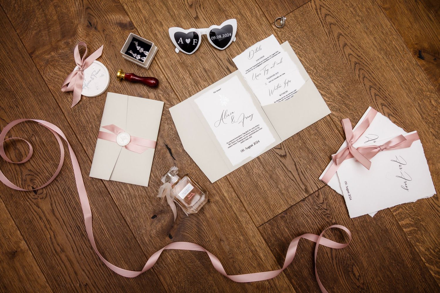 Elegant wedding invitation suite with pink ribbons, perfume bottle, and heart-shaped photo frames on rustic wooden floor.