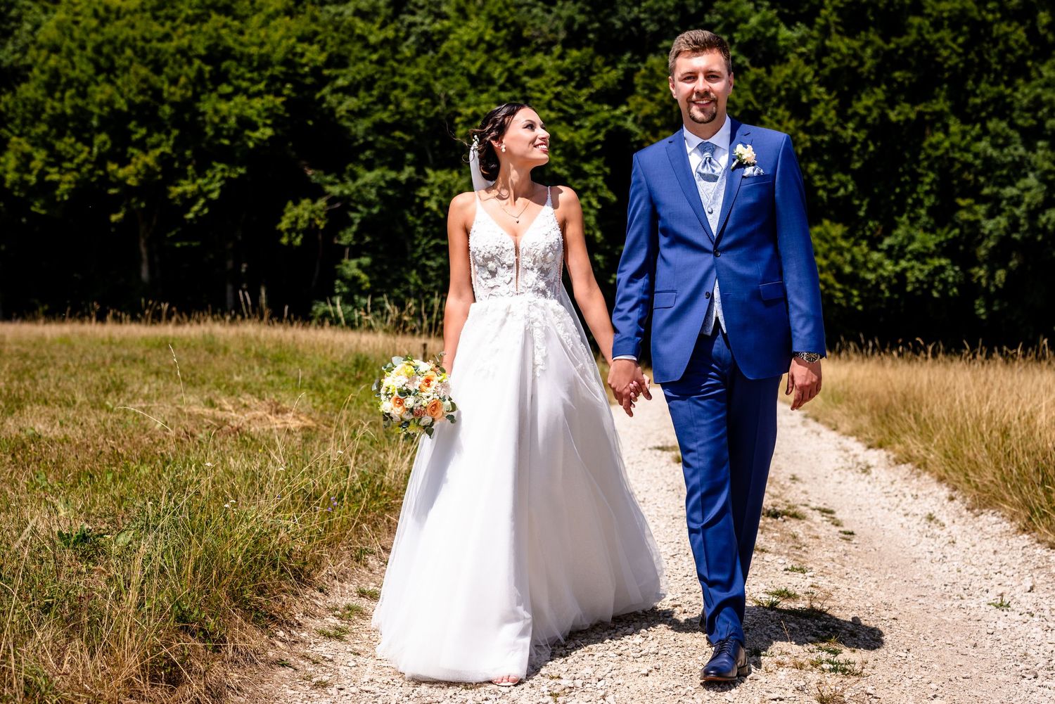 Bride and groom walking hand in hand on a sunny path, she in a white gown, he in a blue suit.