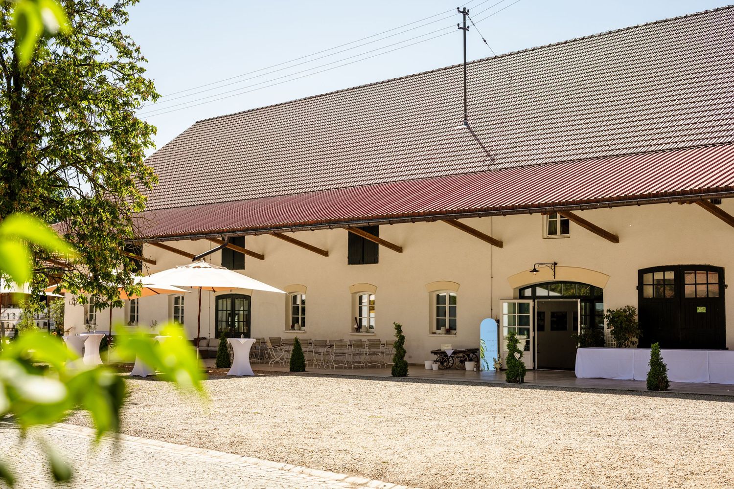 Long beige building with brown roof and multiple windows stretches across a sunny courtyard setting. Fotografiert am Hofgut Sickertshofen