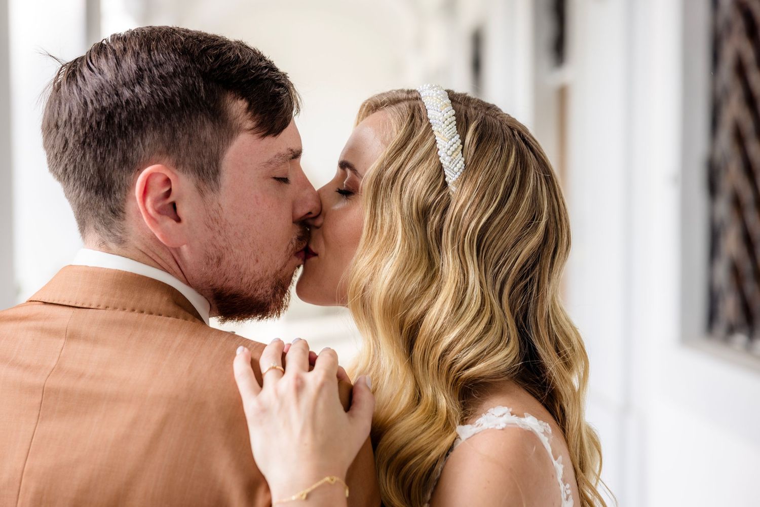 A bride and groom share a romantic kiss, with the bride wearing a lace dress and pearl headband on their wedding day.