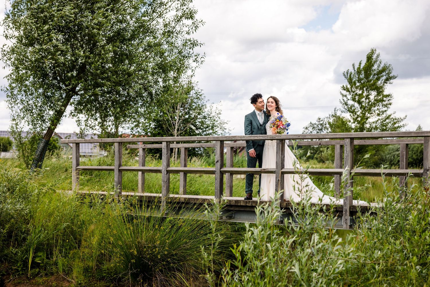 A couple stands together on a wooden footbridge surrounded by lush green summer foliage and wildflowers.