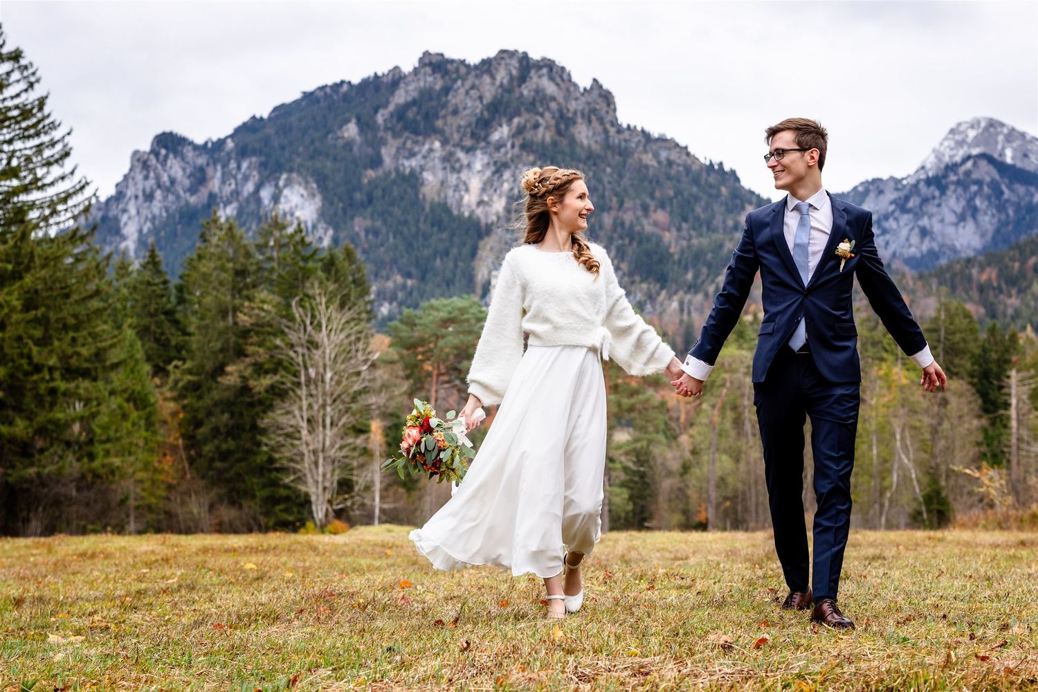 A couple in wedding attire walks hand in hand through a mountain meadow with dramatic peaks rising in the background.