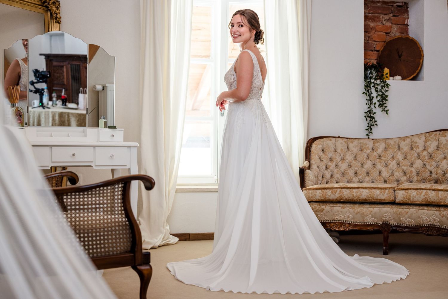 A bride in a flowing white wedding gown stands elegantly in a sunlit room with classic decor and exposed brick walls. Hofgut Sickertshofen