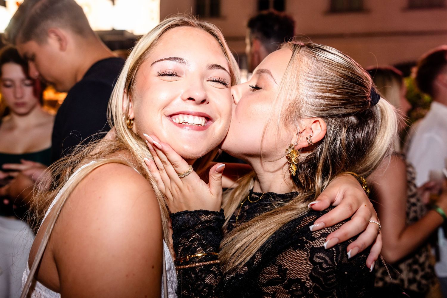 Two blonde women share a joyful moment at a party, one kissing the other on the cheek while smiling.