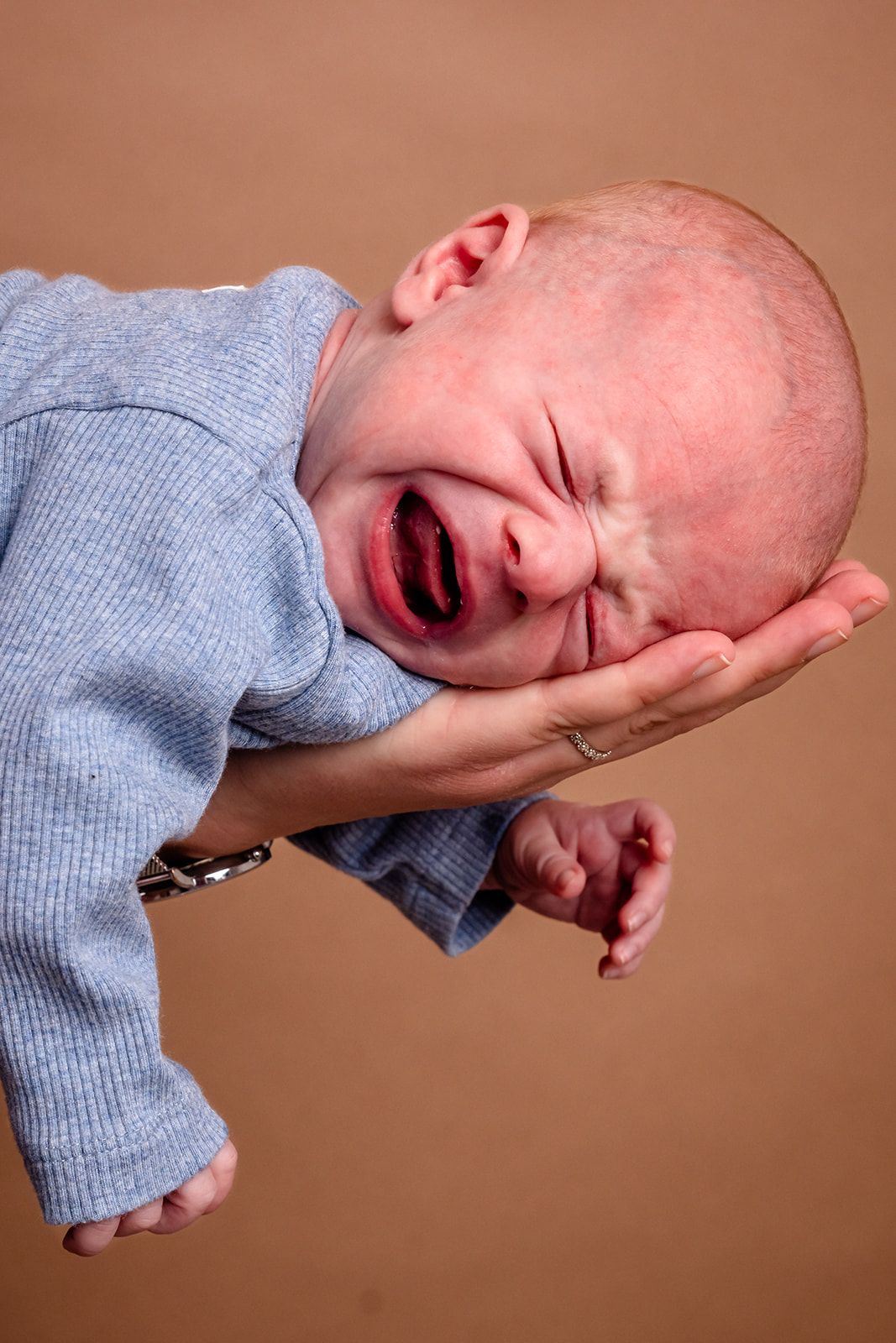 A crying baby in a blue sweater being held up against a brown background.