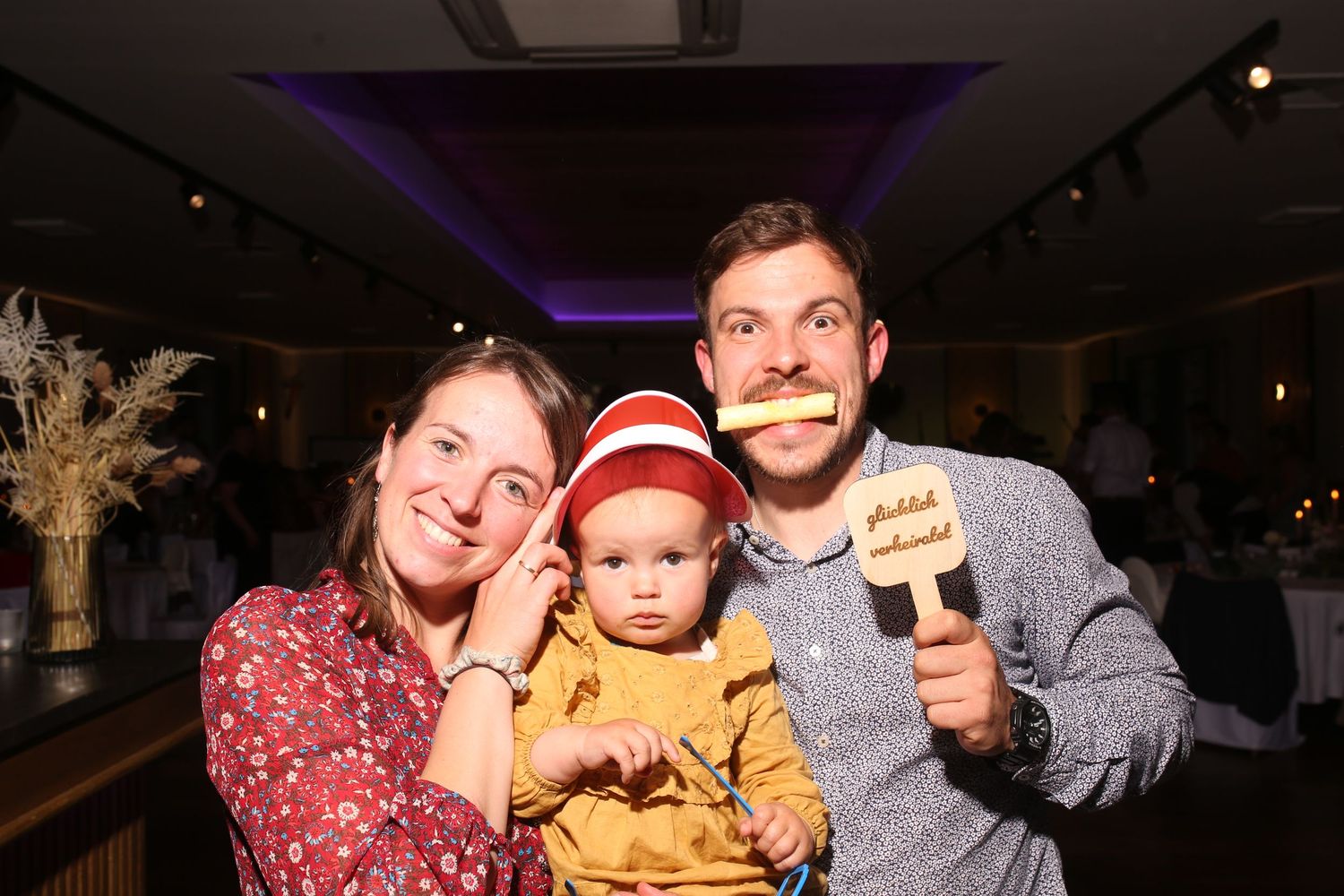 Family of three posing with fun photo booth props at an indoor event, parents holding mustache sticks beside their baby.