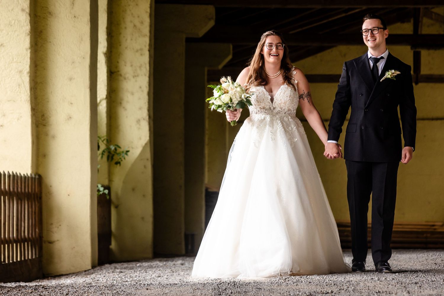 A bride and groom hold hands while walking together, with the bride carrying a white bouquet in her lace wedding gown.