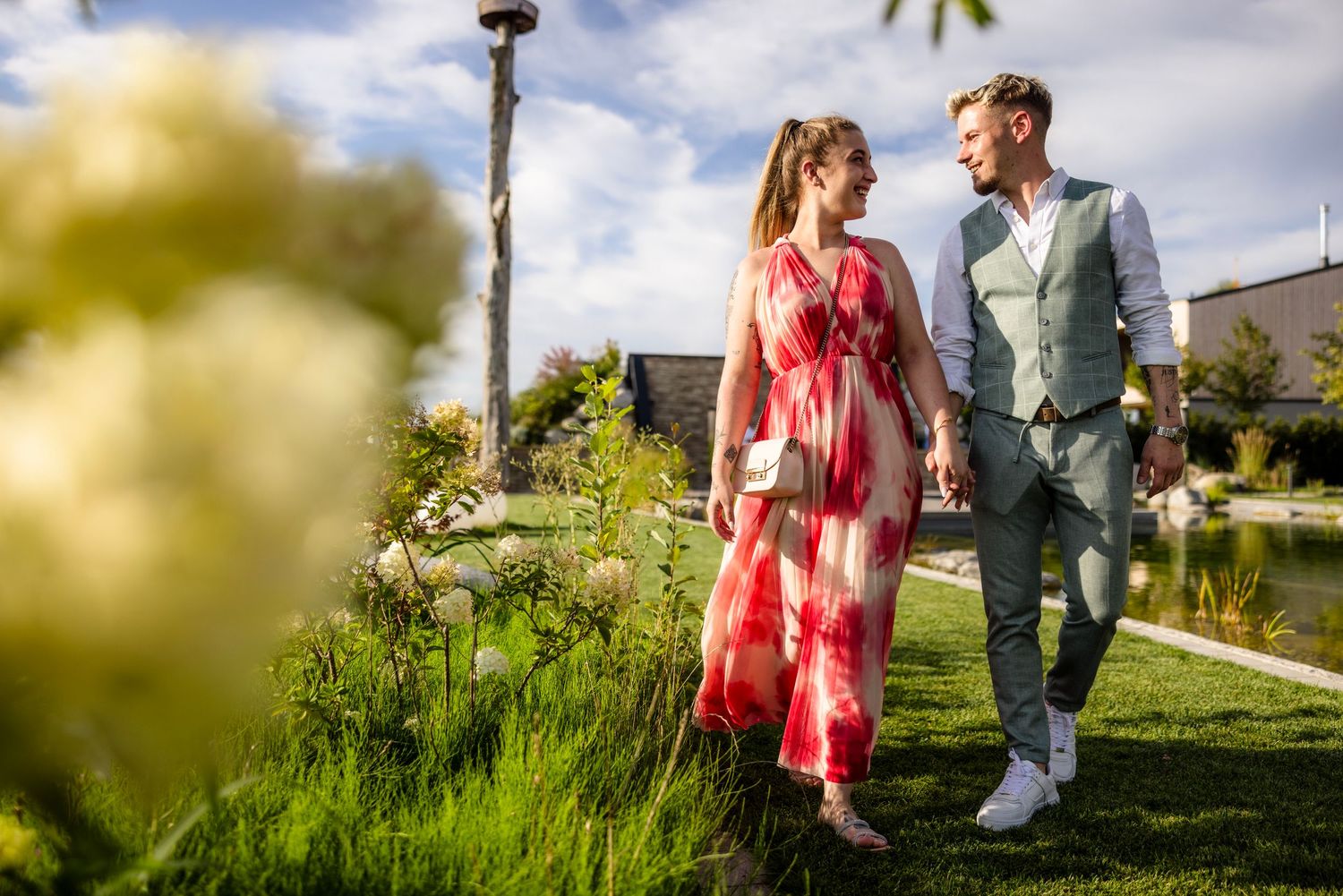 A couple walking together through a lush garden path on a sunny afternoon, wearing casual summer attire.