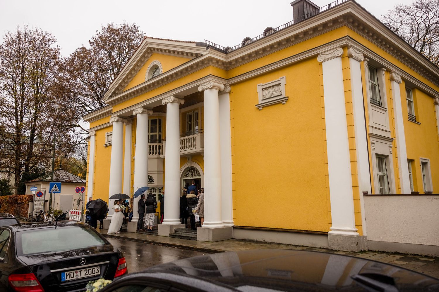 Yellow neoclassical building with white columns on a rainy day, cars parked in front, people with umbrellas at entrance.