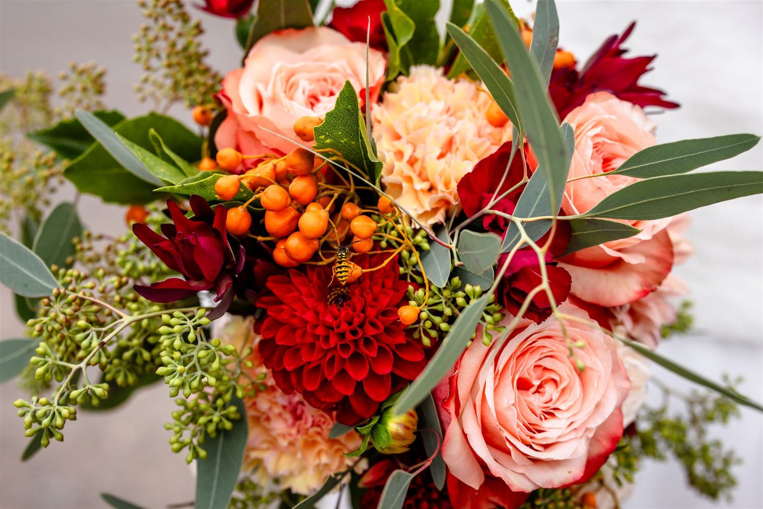 Elegant floral arrangement with coral roses, red dahlias, orange berries and green foliage on white background.
