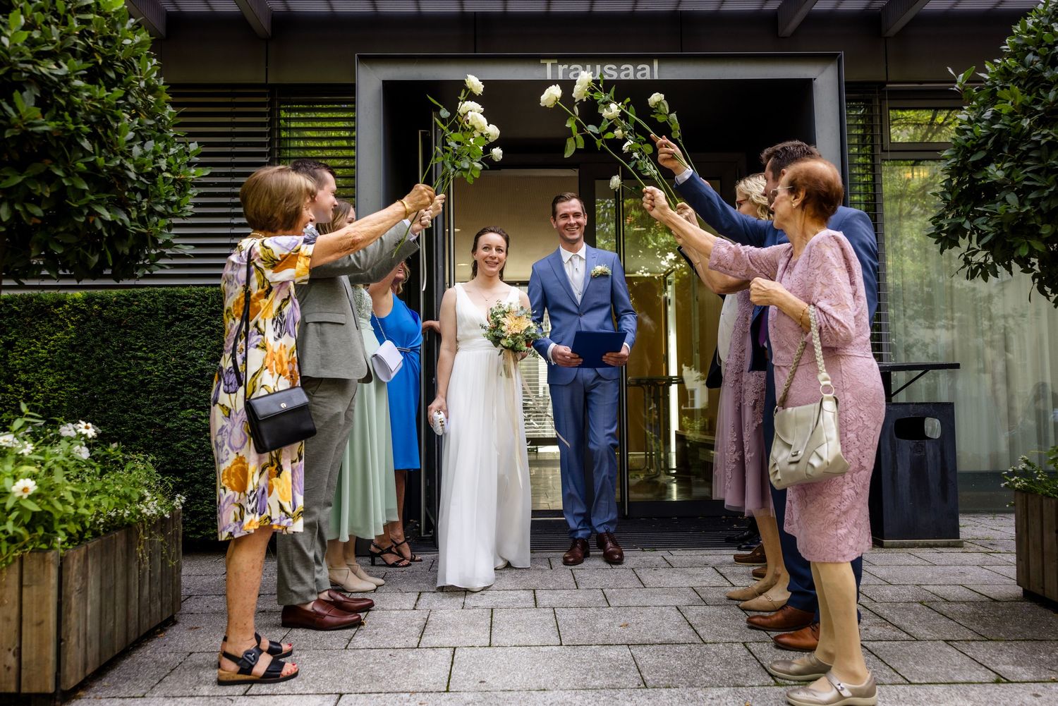 Bride and groom exit venue as guests raise white flowers in celebration at an outdoor wedding ceremony.