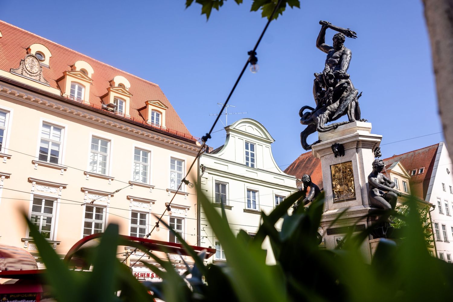 Historic European square featuring a dramatic bronze statue monument with ornate Baroque buildings and clear blue sky in background.