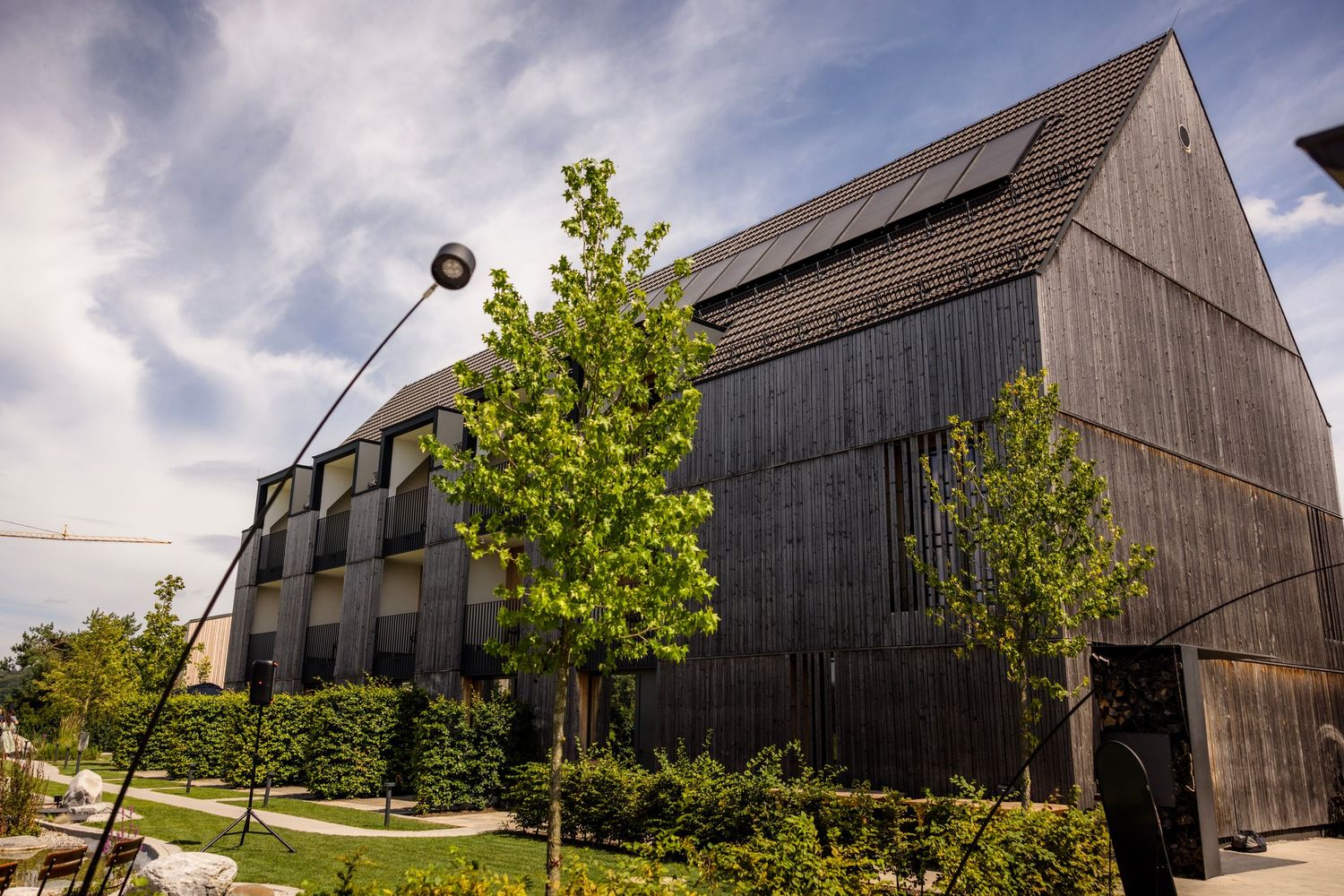 Modern barn-style building with dark wood siding and solar panels on the roof stands among young trees on a sunny day.