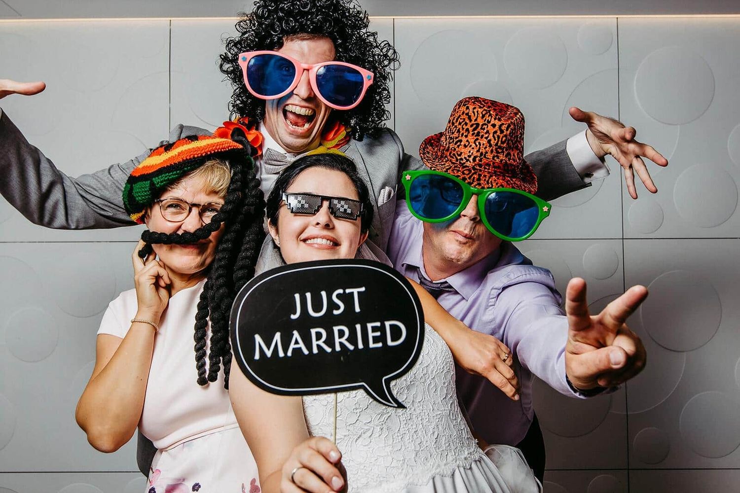 Four people in a photo booth wearing fun props and hats, holding a Just Married sign at a wedding celebration.