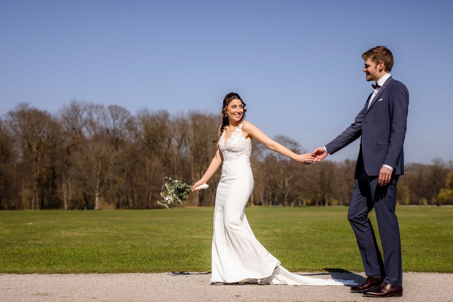 Bride in white gown holds groom's hand in navy suit on sunny day with green park backdrop.