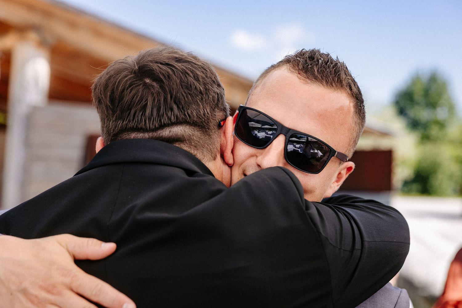Two men in black suits sharing an emotional hug outdoors at what appears to be a wedding celebration.