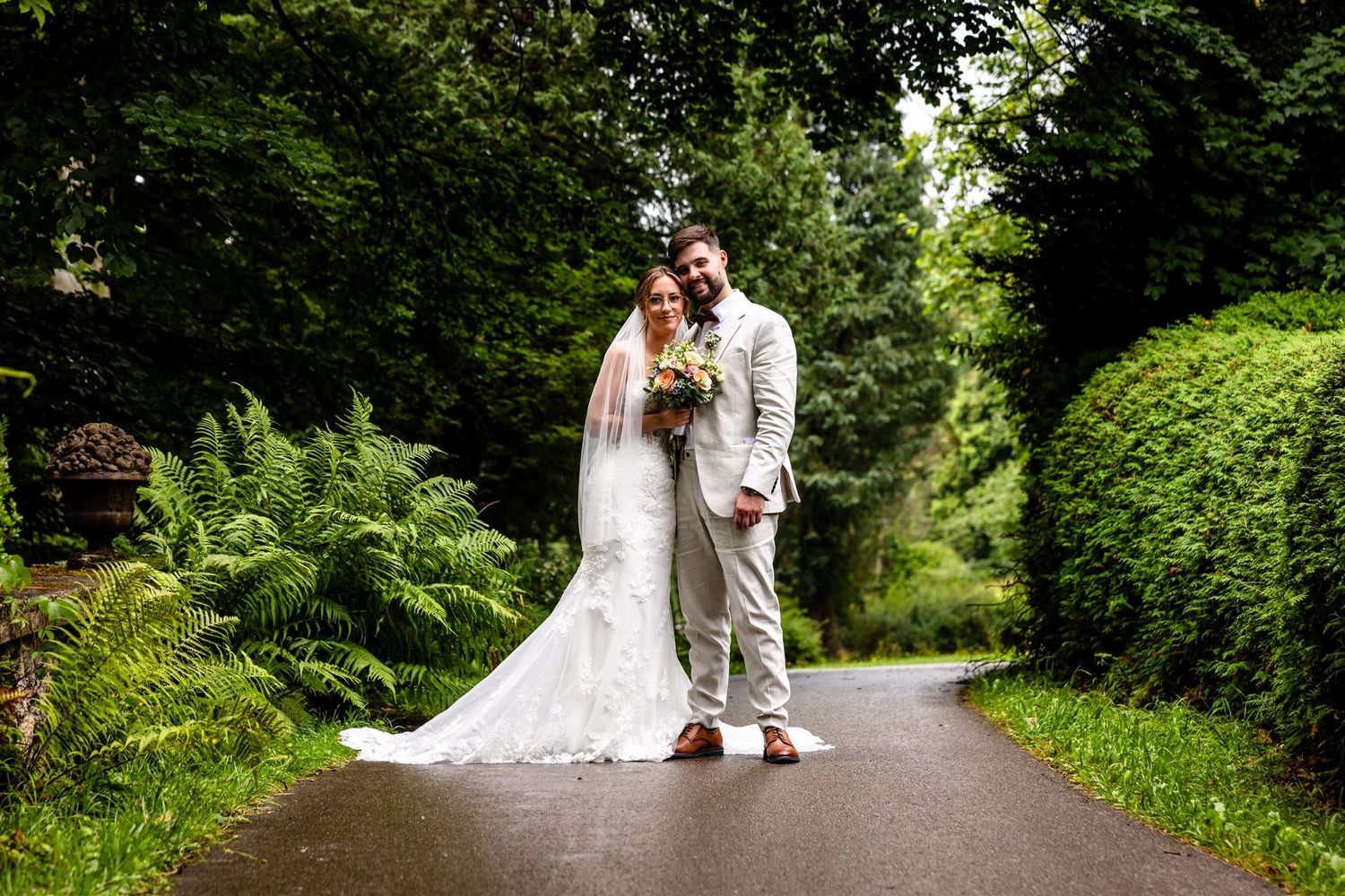 A couple in wedding attire walks down a lush green path surrounded by trees and foliage.