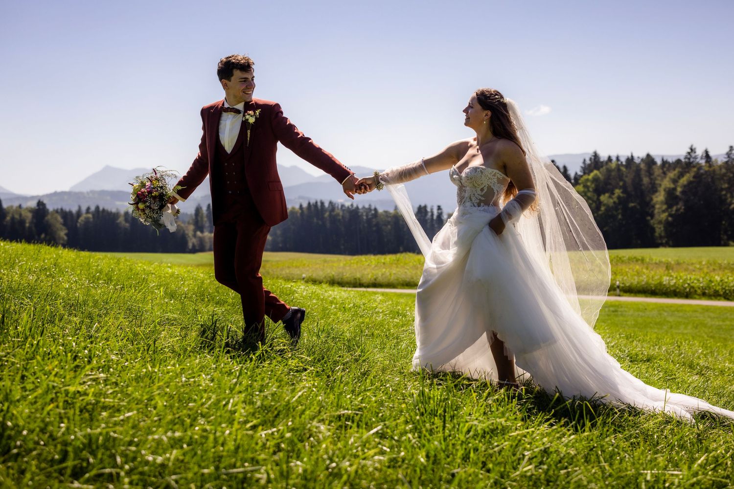 Bride and groom holding hands walking through a lush green meadow with mountain views on a sunny wedding day.