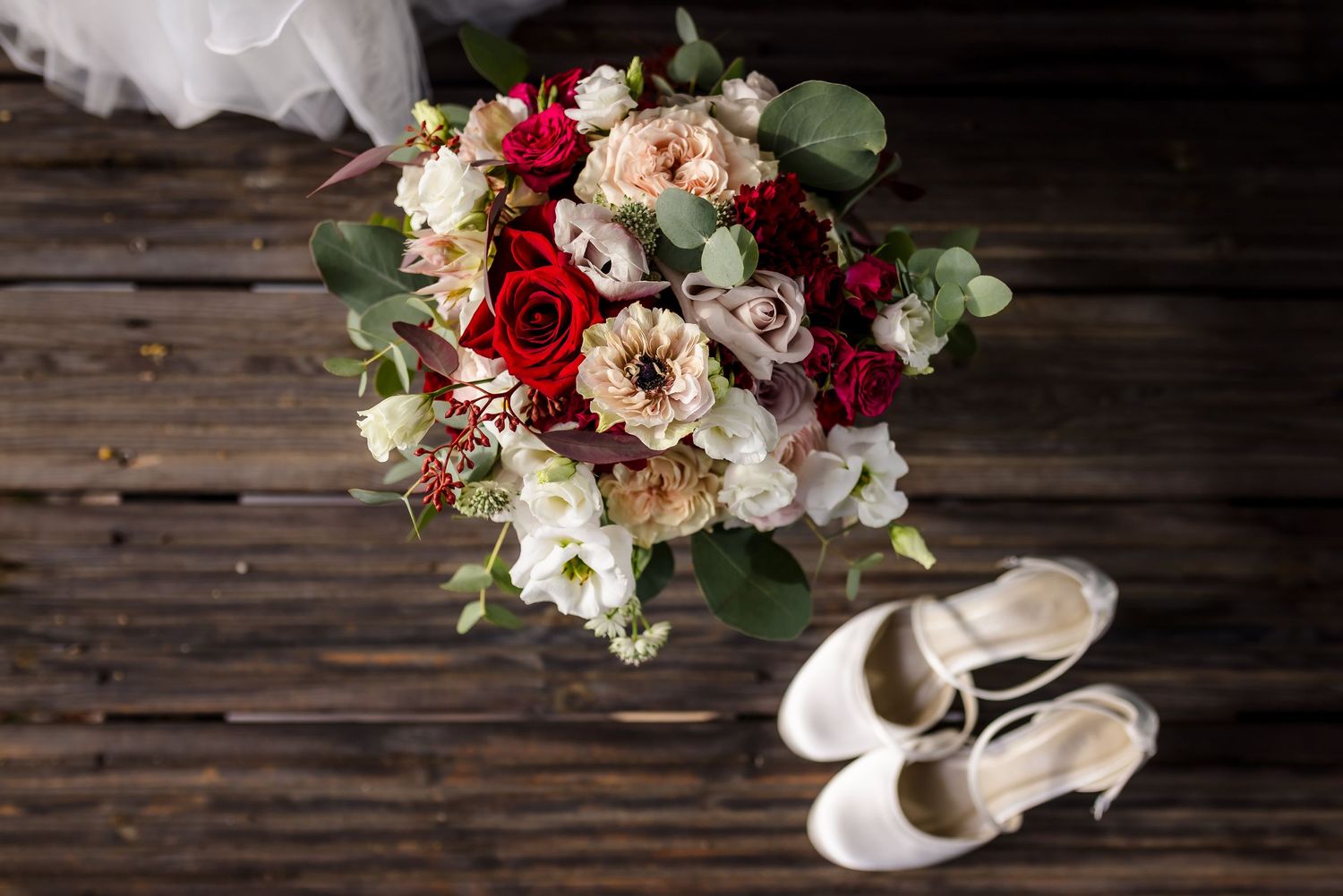 Brautstrauß mit roten und cremefarbenen Rosen neben weißen Hochzeitsschuhen auf einem rustikalen Holzboden. Preise für Hochzeitsfotografen