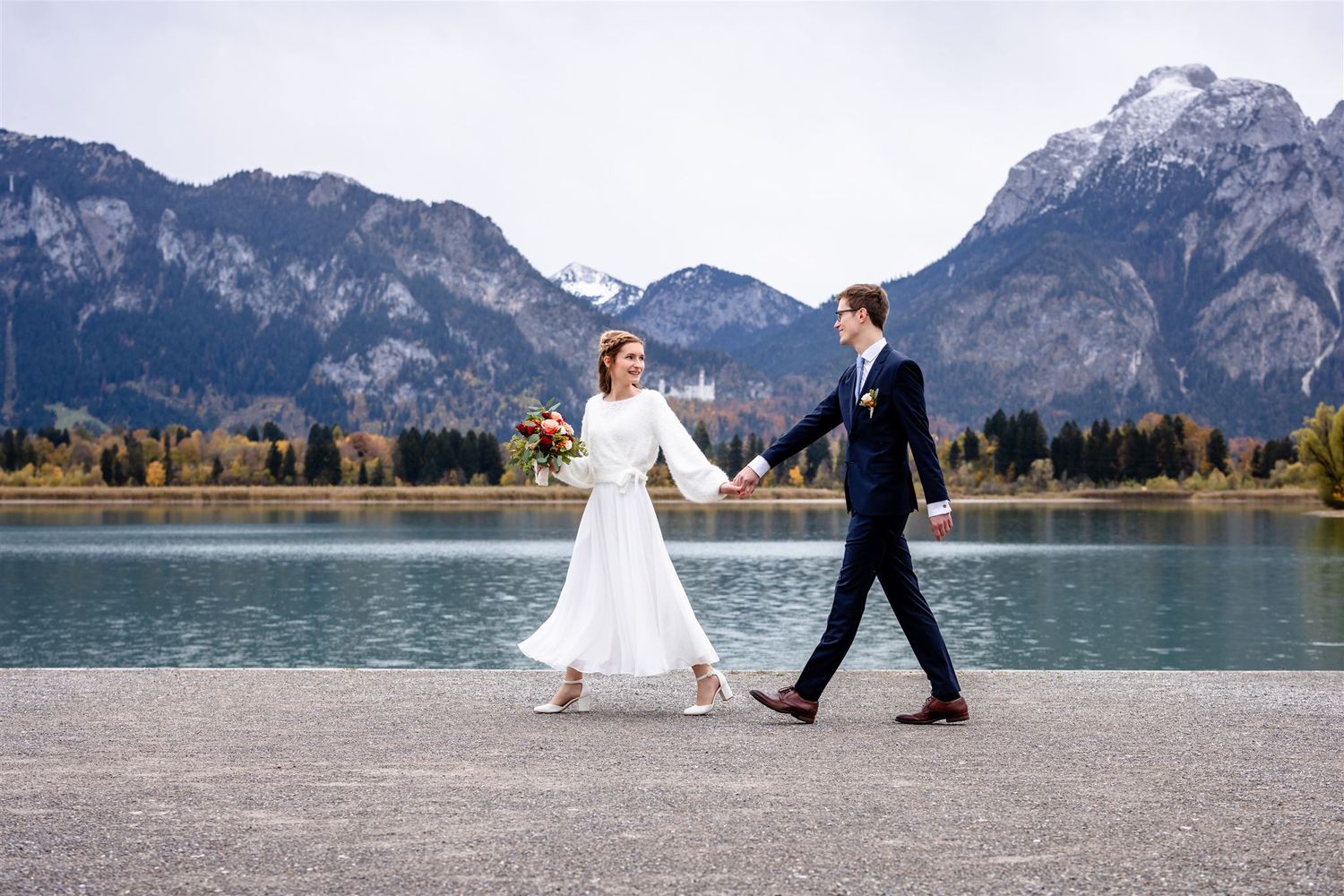 A couple in wedding attire walks along a scenic mountain lake with snow-capped peaks in the background.