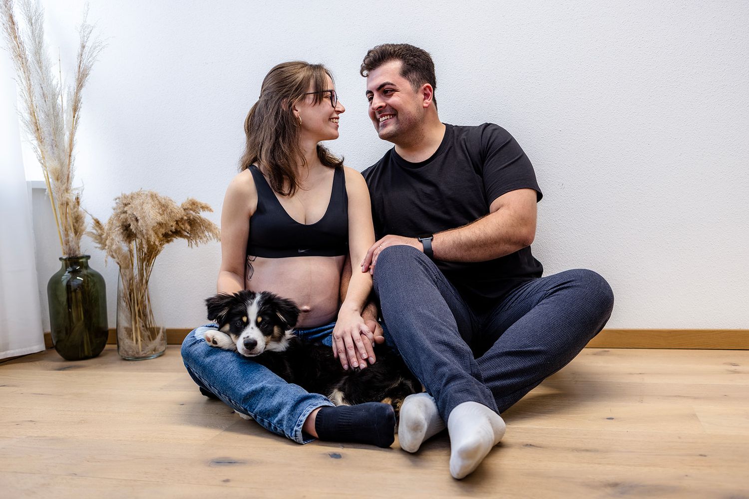Two people sit together on a wooden floor in a minimalist room with white walls and dried pampas grass decor.