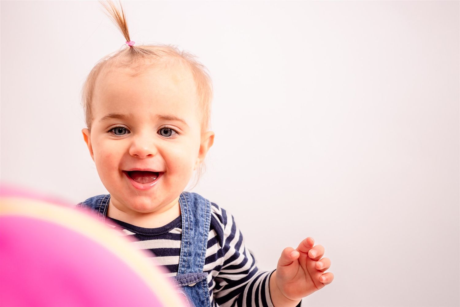 Baby in a navy striped outfit smiles and plays while sitting on a pink background.