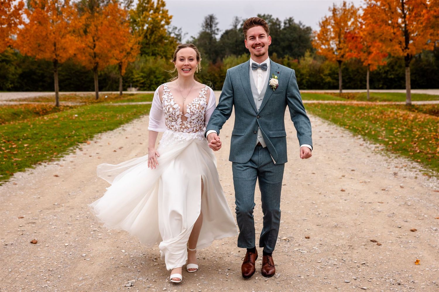 A couple walks down a gravel path surrounded by vibrant orange autumn trees in a romantic wedding photo shoot.