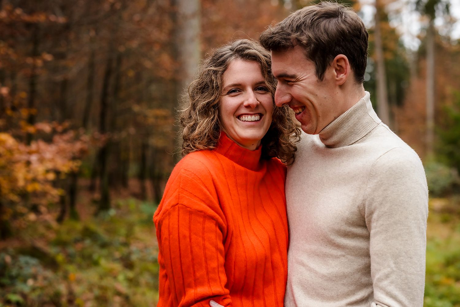 A couple wearing orange and beige sweaters share intimate moments while walking through an autumn forest setting.