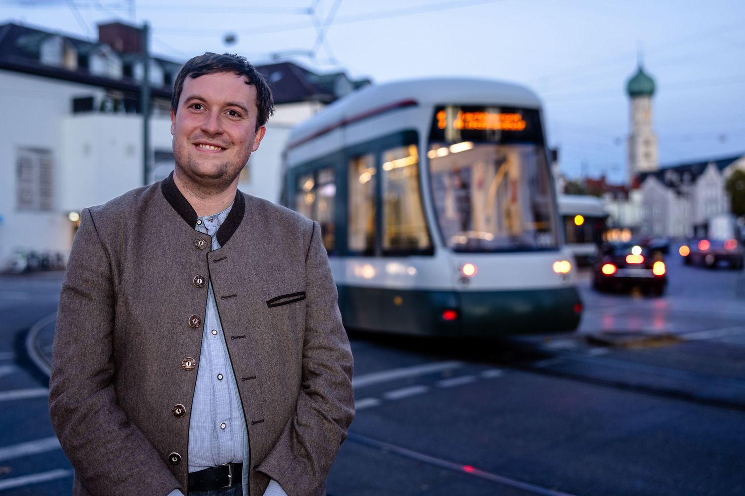 Man in traditional Bavarian jacket smiling in front of a tram on a city street at dusk.