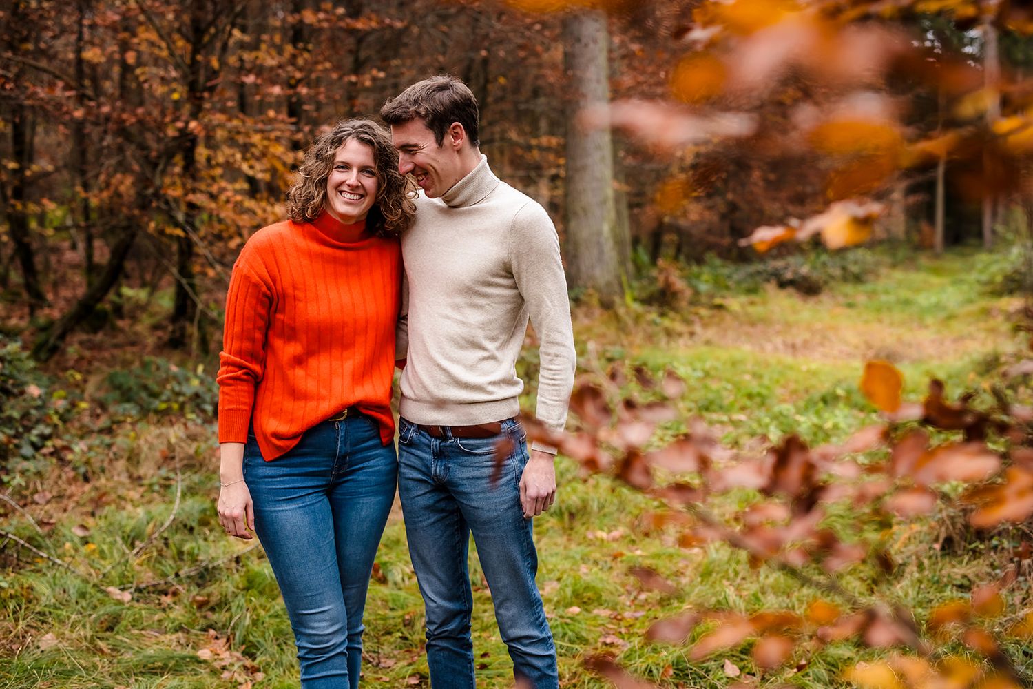 A couple in autumn attire walks together through a forest with orange falling leaves and foliage.