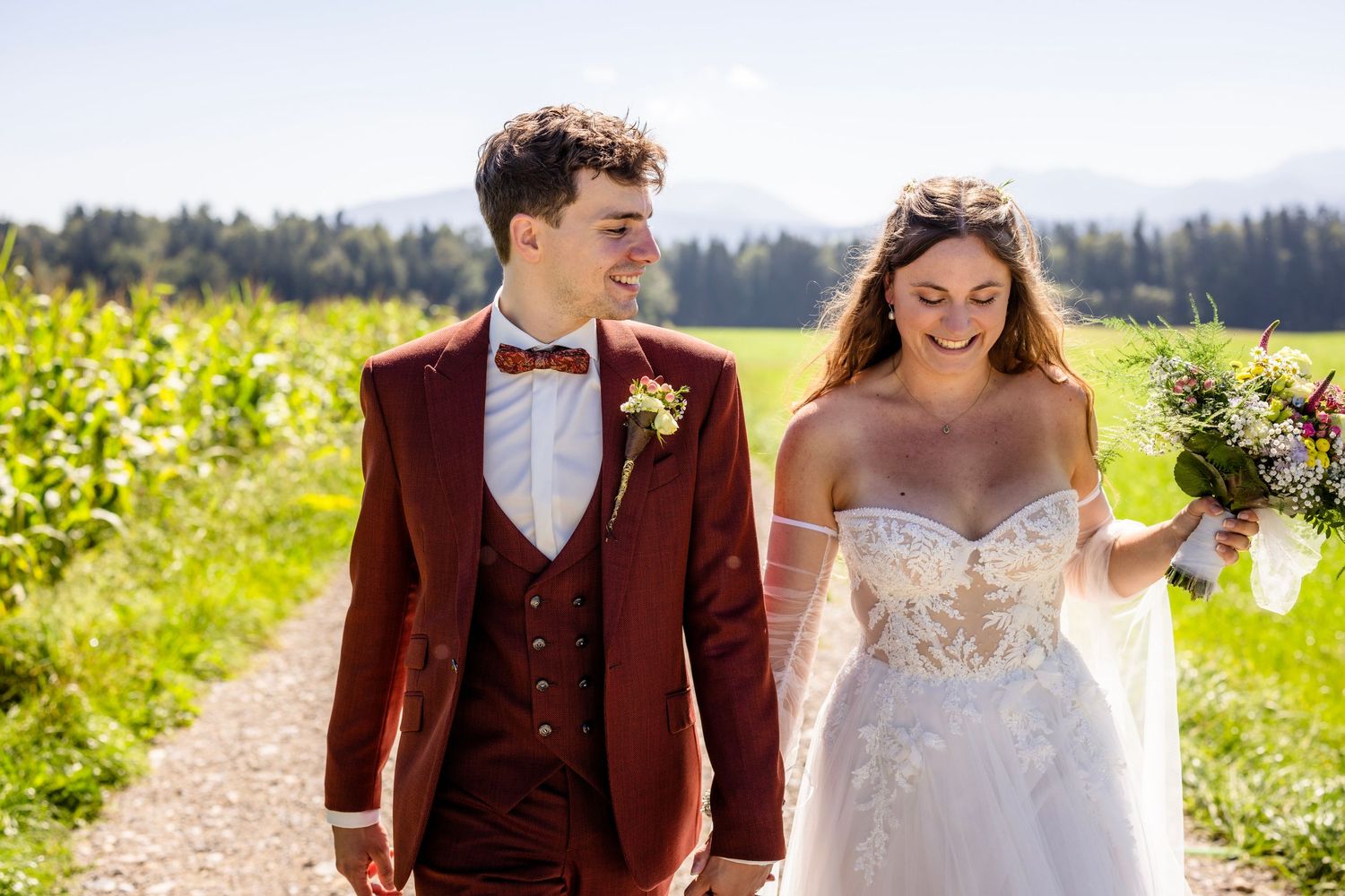 A smiling bride and groom walk outdoors on their wedding day, with mountains and green fields in the background.