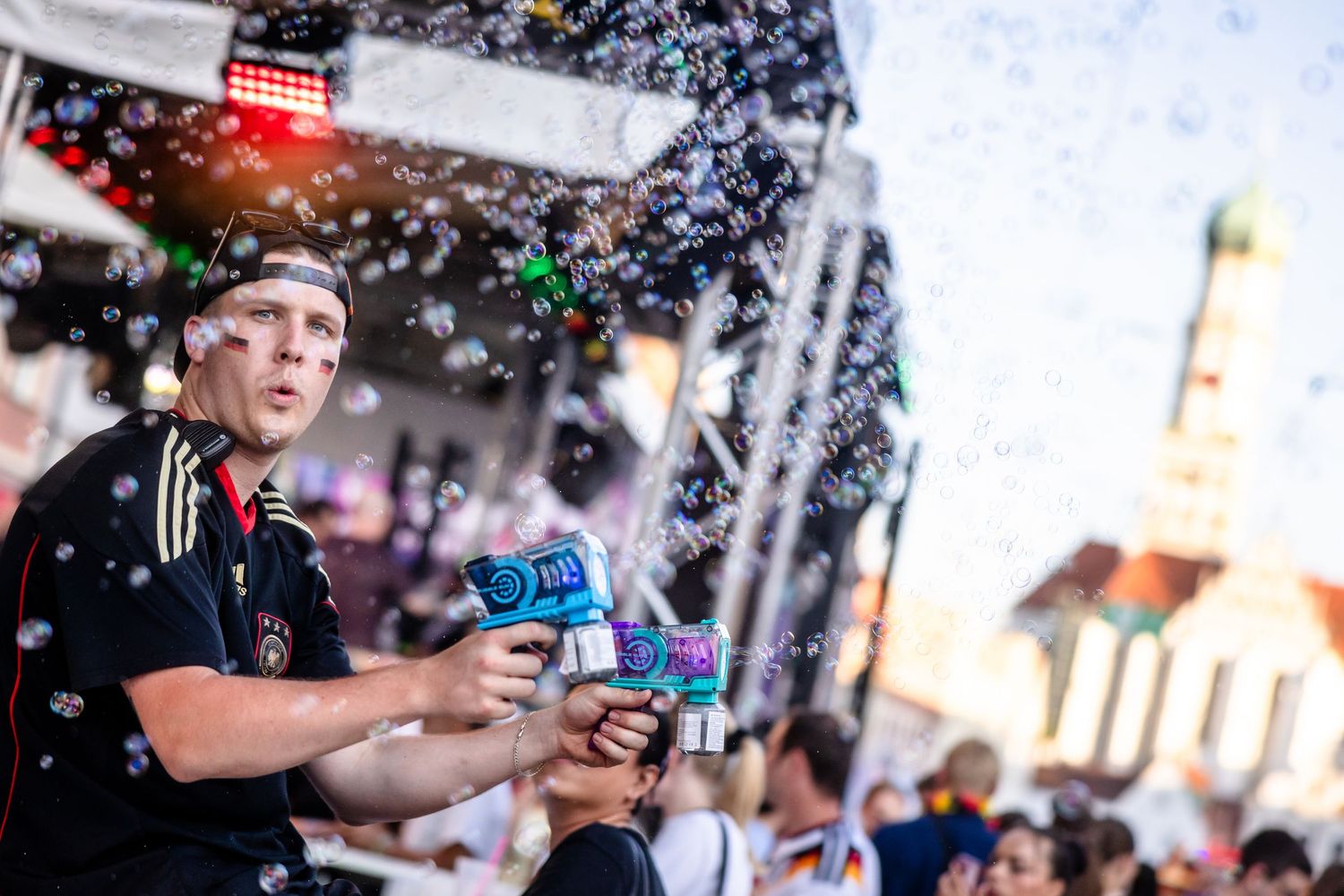 Man in German jersey shoots bubble gun at lively outdoor festival with confetti and crowds in background.