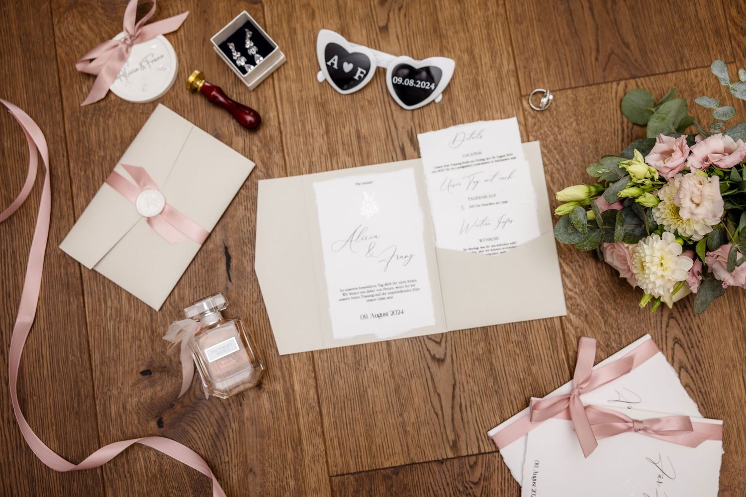 Elegant wedding stationery flat lay with pink ribbons, floral bouquet, perfume bottle, and decorative accessories on wood.