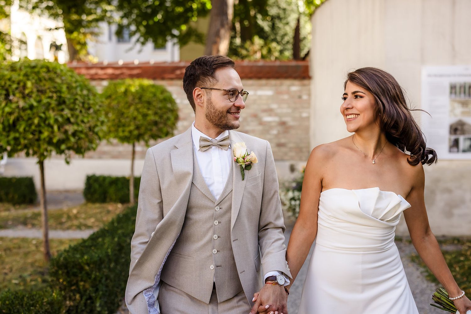 Happy newlyweds share a joyful moment while walking together in a garden setting wearing formal wedding attire.