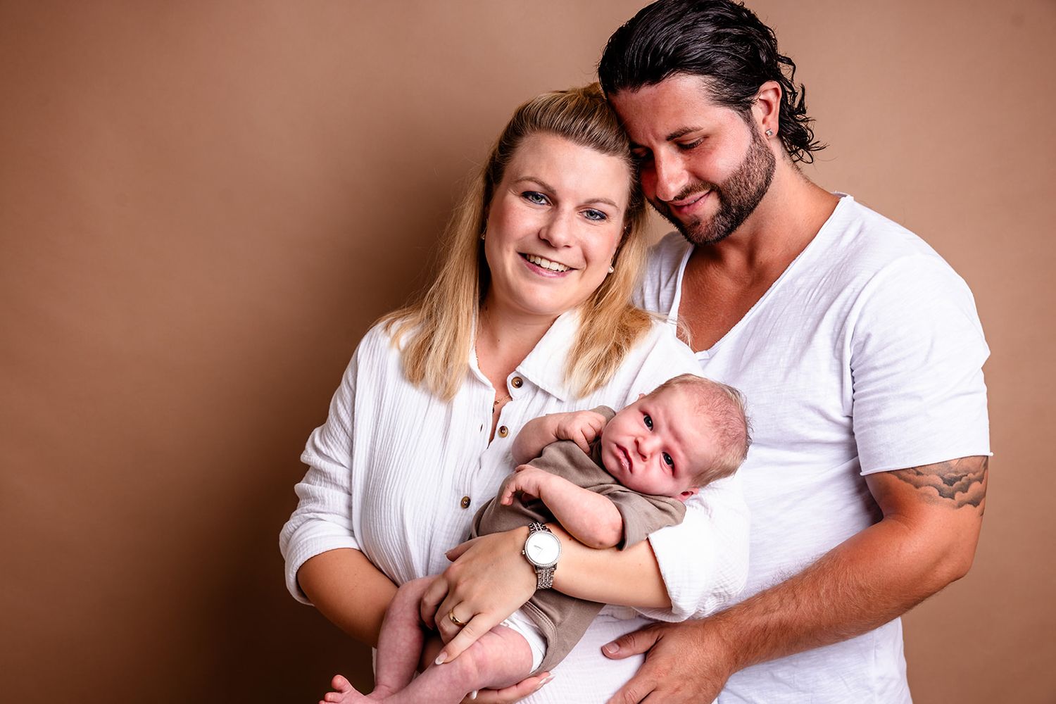 A series of tender family portraits showing parents cradling their newborn baby in matching white outfits against a brown backdrop.