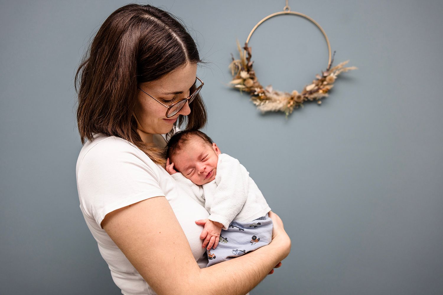 A tender moment between parent and newborn baby against a gray wall with a decorative floral hoop hanging.