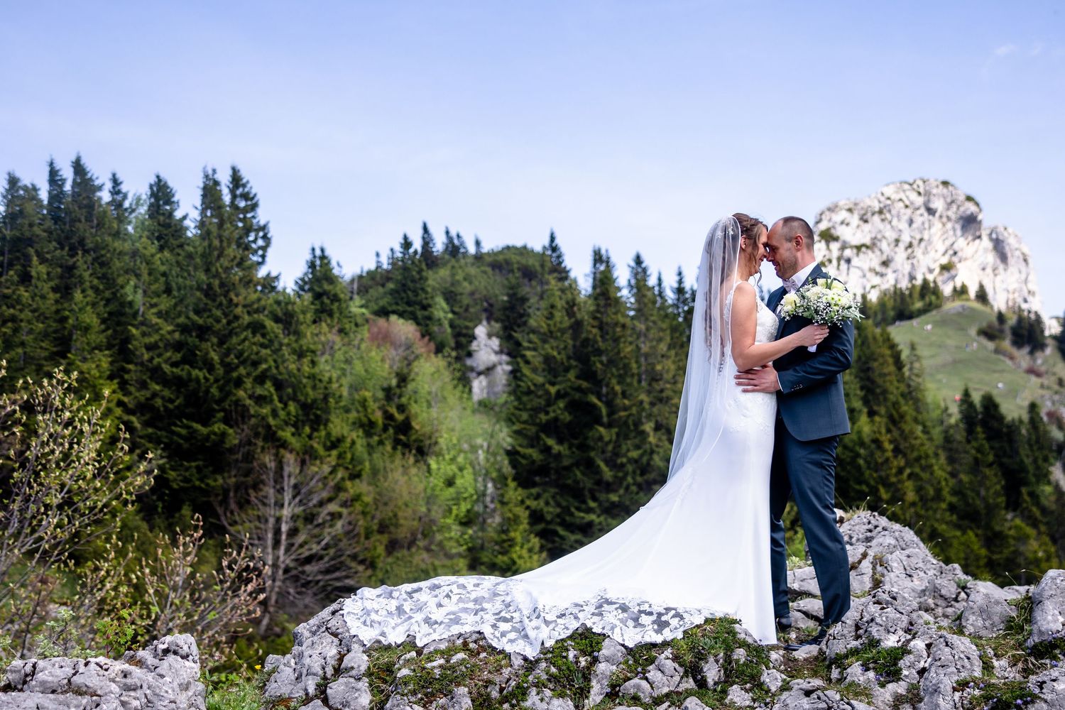 A bride and groom share a romantic kiss on rocky terrain with lush pine trees and a mountain peak backdrop.